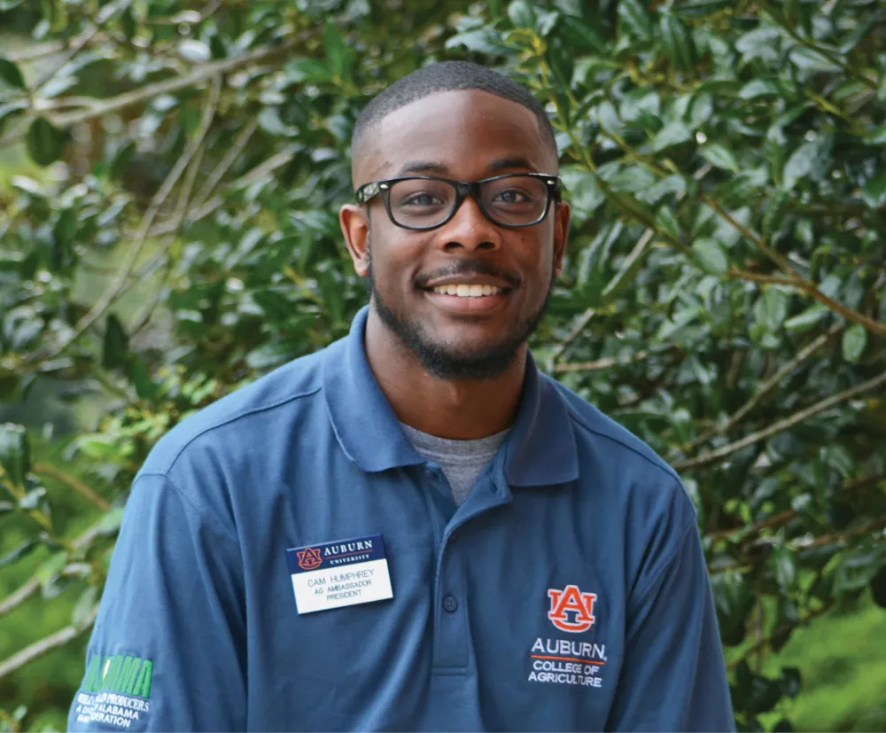 Portrait of a smiling man wearing an Auburn College of Agriculture polo shirt with a name tag reading "Cam Humphrey, Ag Ambassador President," standing in front of green foliage.