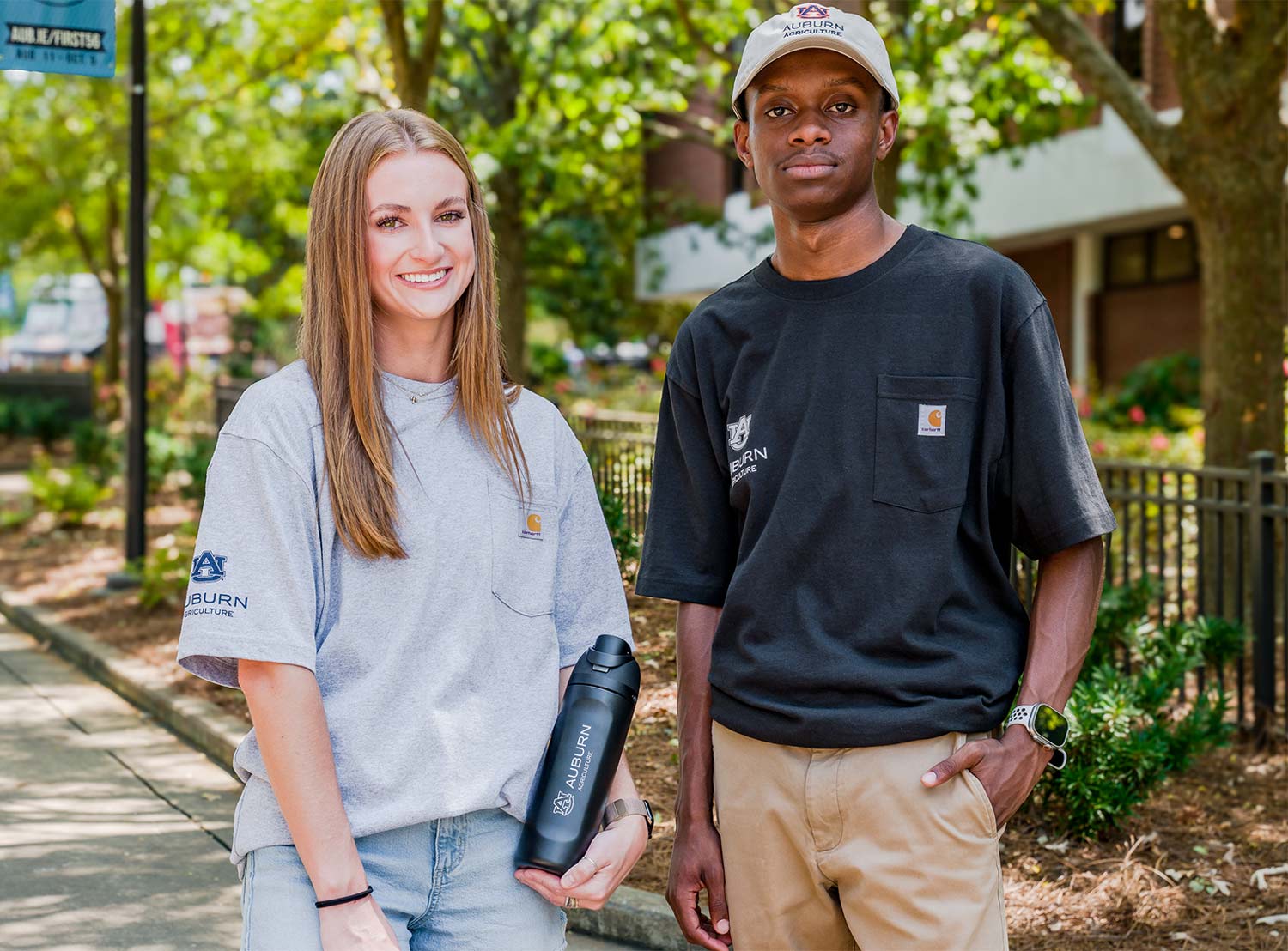 A man and woman wear Auburn Agriculture shirts and a hat while standing on a sunny campus walkway.