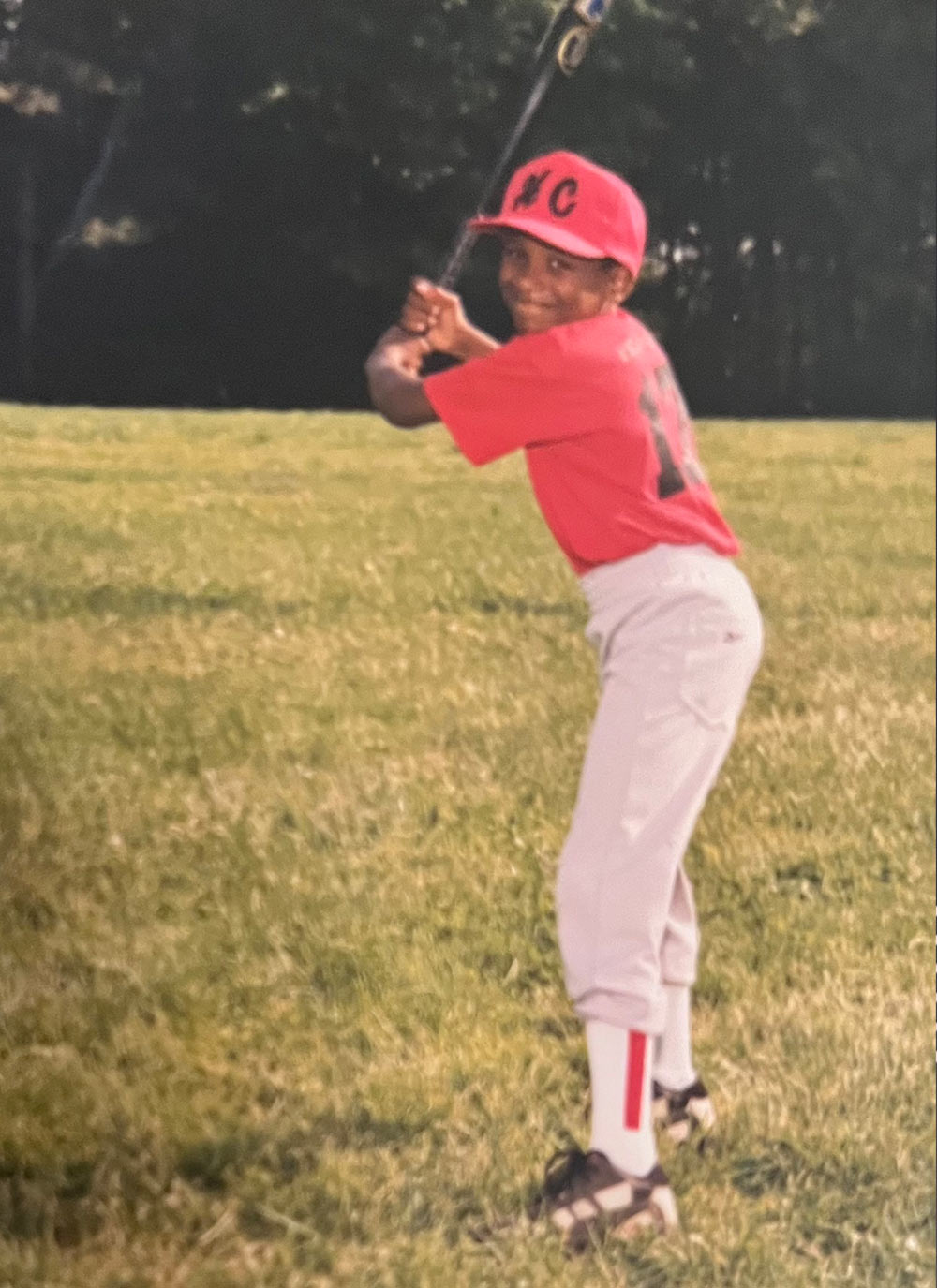 A young Blake Poole in a red baseball jersey and cap holds a bat over his shoulder, prepared to swing. He stands on a grassy field with a line of trees in the background, smiling toward the camera.