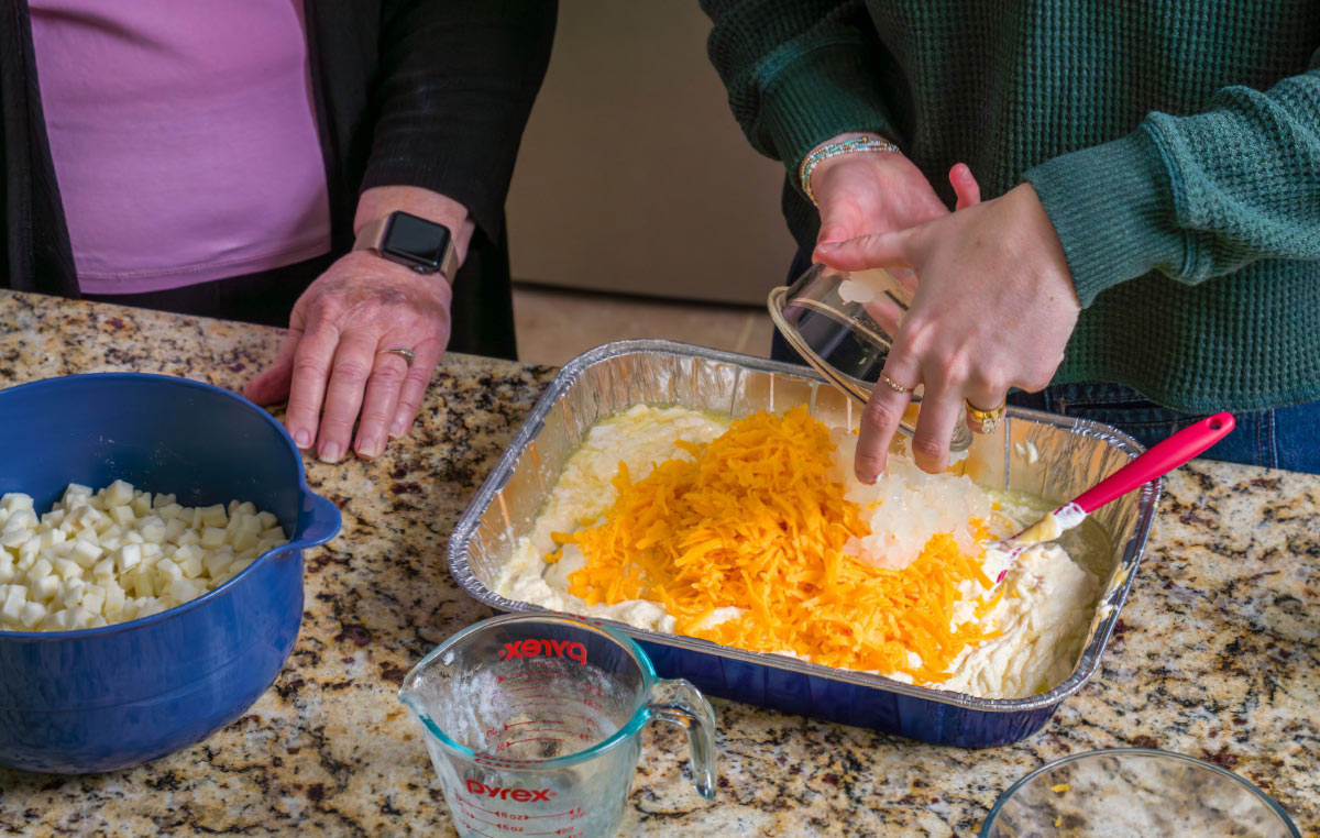 A young woman in a green sweater pours shredded cheese and diced onions into a foil pan filled with a creamy potato mixture.