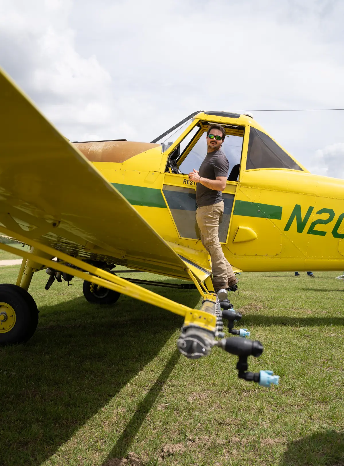 A man wearing sunglasses stands on the wing of a yellow aircraft, giving a thumbs-up gesture; The plane has a spray boom with blue nozzles extending along the wing, and its registration number begins with N20