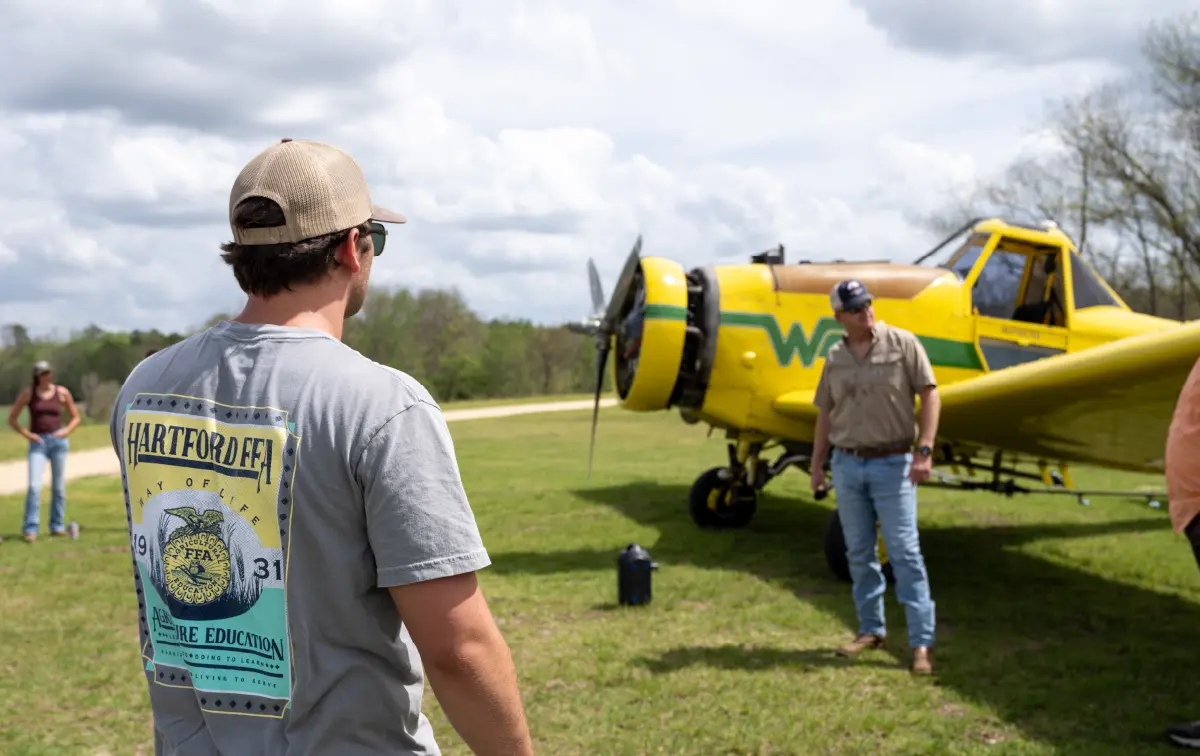A young man wearing a grey graphic t-shirt that says Hartford FFA Agriculture Education on it and a tan beige hat stands in a field looking toward a yellow aircraft; A man in a tan button-up dress shirt, blue denim jeans, a hat, and sunglasses stands near the plane's wing