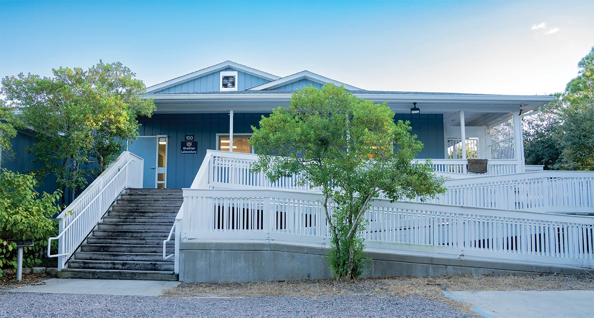 Exterior view of a blue coastal-style building with white railings and an accessible ramp, surrounded by trees and gravel, identified as a laboratory or facility entrance.