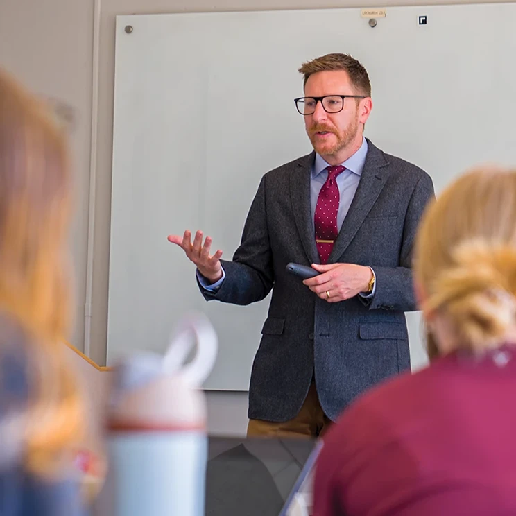 A male instructor stands at the front of a classroom, gesturing while speaking to seated students, with a whiteboard behind him and students partially visible in the foreground.