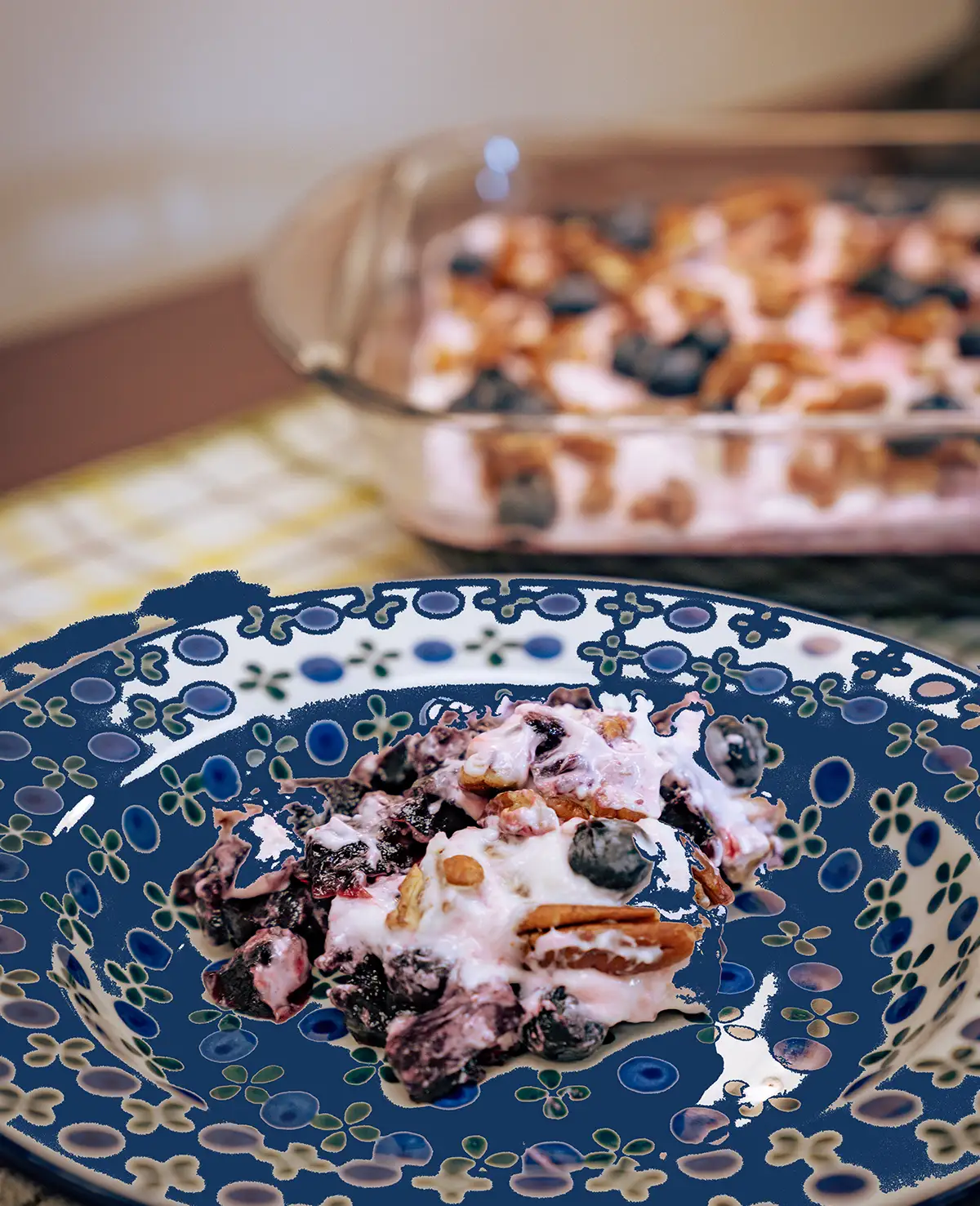 A serving of a creamy berry dessert mixed with blueberries and pecans on a blue patterned plate, with the full glass baking dish blurred in the background.