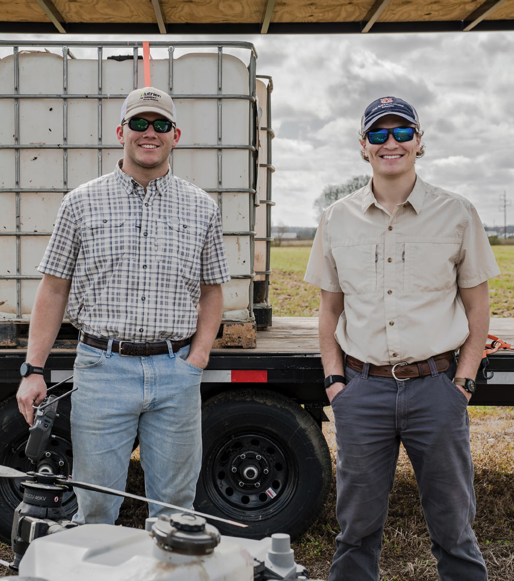 Two smiling men wearing sunglasses and baseball caps stand in a field next to a trailer with agricultural equipment.