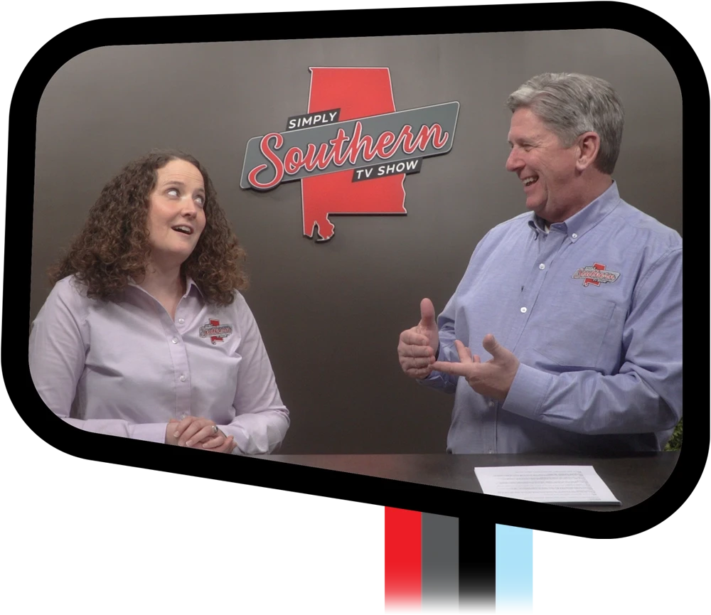 A woman with curly hair and a man with grey hair laugh and gesture while hosting on a TV set. Behind them is a grey wall featuring a red Alabama state outline and the "Simply Southern TV Show" logo.